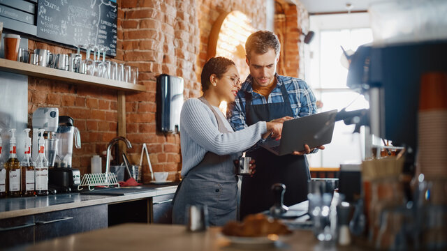 Two Diverse Entrepreneurs Have A Team Meeting In Their Stylish Coffee Shop. Barista And Cafe Owner Discuss Work Schedule And Menu On Laptop Computer. Multiethnic Female And Male Restaurant Employees.