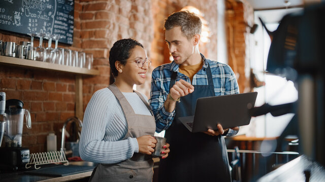 Two Diverse Entrepreneurs Have A Team Meeting In Their Stylish Coffee Shop. Barista And Cafe Owner Discuss Work Schedule And Menu On Laptop Computer. Multiethnic Female And Male Restaurant Employees.