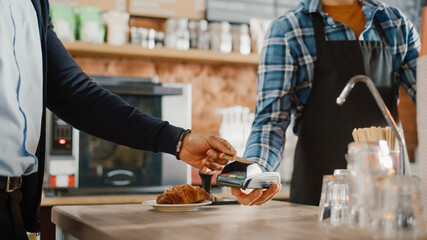 African American Customer Pays for Coffee and Pastry with Contactless NFC Payment Technology on Smartphone to a Handsome Barista in Blue Checkered Shirt. Contactless Mobile Payment in Cafe Concept.