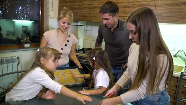 Mom holding baking tray and daughters putting raw gingerbread cookies on it, father helping. Home video of happy family cooking together before Christmas. Concept of holiday