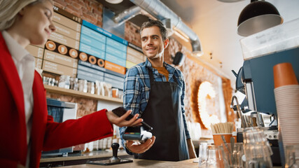 Female Customer Pays for Take Away Coffee with Contactless NFC Payment Technology on Smartphone to a Handsome Barista in Checkered Shirt in Cafe. Customer Uses Mobile to Pay Through Bank Terminal.