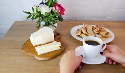 Cup of coffee in female hands, close-up. Homemade baked goods during quarantine. Homemade feta cheese on a wooden tabletop. Concept on the background of a white brick wall and a bouquet of roses.