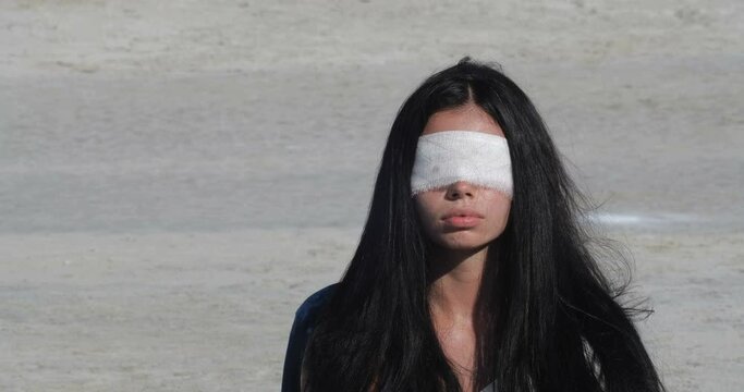 Close up  portrait of a woman with a blindfold and long black hair. Blood stains appear on the bandage. Outdoors desert shooting. 4K fps slow motion