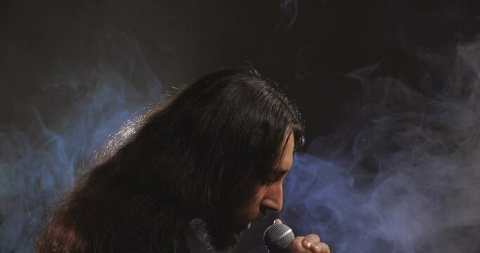 Dramatic close-up portrait of a long-haired Caucasian male, brunet with a beard against a black background with smoke, wind and backlight. Rock vocalist screaming to microphone. 50 fps 4k