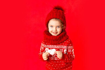 Fototapeta premium a little girl in winter clothes holds a heart on a red background. New year's or Valentine's day concept, place for text