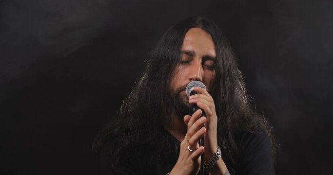 Dramatic close-up portrait of a long-haired Caucasian male, brunet with a beard against a black background with smoke, wind and backlight. Rock vocalist screaming to microphone. 50 fps 4k