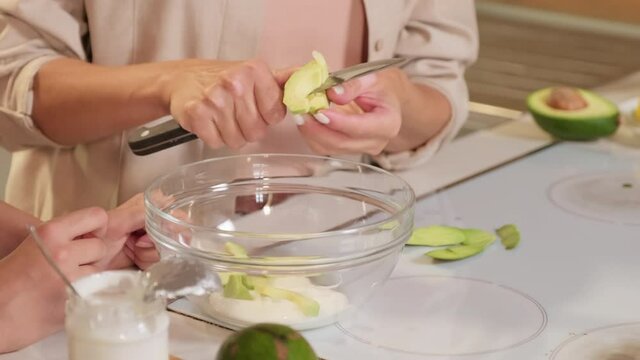 Slow-motion closeup of unrecognizable female hands cutting ripe peeled avocado in small slices to bowl preparing ingredients for homemade healthy ice cream