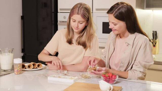 Medium footage of happy mom and her teenage daughter standing at kitchen table decorating ice-cream with nits and fruit laughing and having fun together
