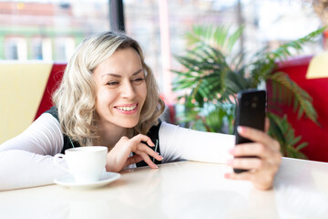 Online date. Smiling and laughing adult caucasian woman chatting in cafe during a video call, smartphone in hand, selective focus.