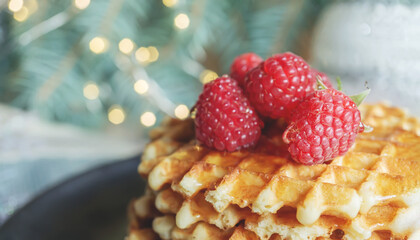 Viennese waffles with raspberries and honey on the Christmas table with tree branches and garlands of lights in the background. close-up, copy space