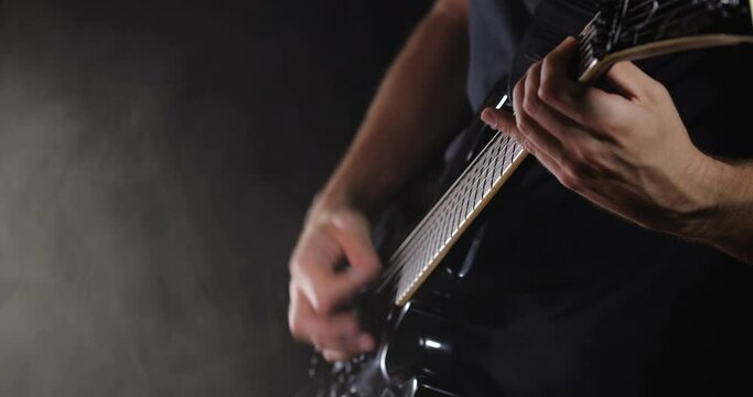 Unrecognizable rock musician in black clothes plays black electric guitar on black background isolated and smoke. 