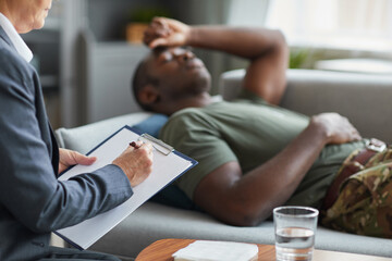 Close-up of woman writing a prescription in her document with her patient lying on sofa in the background