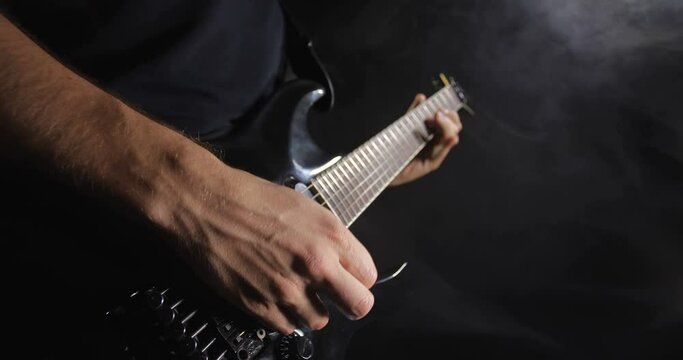 Unrecognizable rock musician in black clothes plays black electric guitar on black background isolated and smoke. 