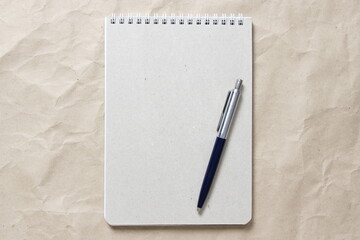 Gray notepad with white coiled spring and pen on a background of beige crumpled craft paper. With empty space for text and design