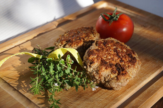 zwei hausgemachte Frikadellen, Bouletten, Fleischpflanzerl auf einem Holzbrett mit Bund von frischen Kr&auml;utern und Tomate