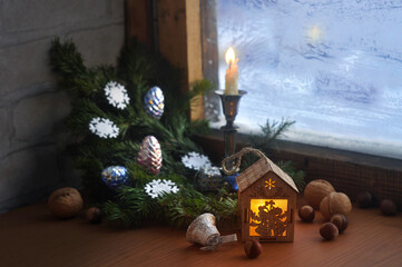 Wooden toy house, burning candle, fir branches and Christmas toys on a snowy wooden window. Winter evening.