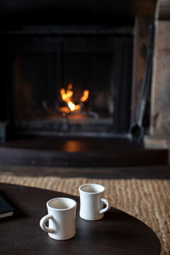 Two mugs of coffee on a table in front of wood burning fireplace