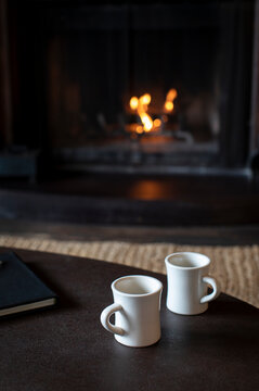 Two mugs of coffee on a table in front of wood burning fireplace