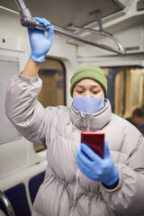 Fototapeta na wymiar Young woman in protective gloves and mask using mobile phone while standing on a train