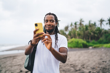 Cheerful black man surfing cellphone on tropical coastline