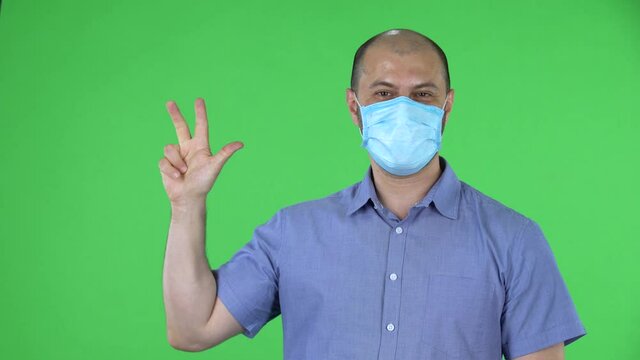 Portrait of a middle aged men in medical mask shows fist, then counting one, two, three, four, five fingers. Balding male in blue shirt posing on green screen in the studio. Close up.