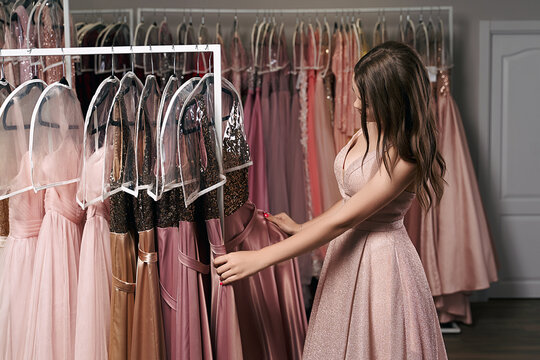 Portrait of young beautiful girl wearing a full-length pale pink glitter chiffon draped prom ball gown. Model selecting an outfit for occasion in dress hire service with many options on background.