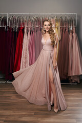 Young beautiful blonde girl wearing a full-length pale pink glitter chiffon draped prom ball gown. Model selecting an outfit for occasion in dress hire service with many options on background.