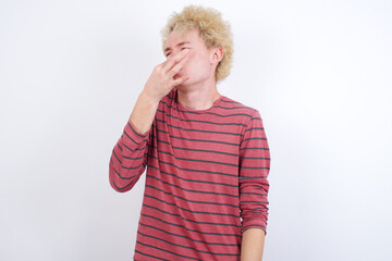 Young handsome Caucasian blond man standing against white background smelling something stinky and disgusting, intolerable smell, holding breath with fingers on nose. Bad smell