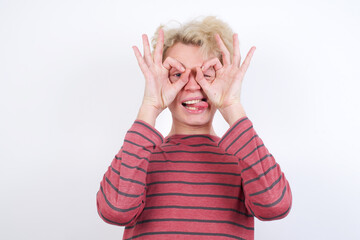 Young handsome Caucasian blond man standing against white background doing ok gesture like binoculars sticking tongue out, eyes looking through fingers. Crazy expression.