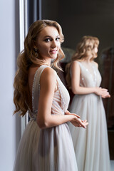 Young beautiful blonde girl wearing a full-length silver white chiffon prom ball gown decorated with sparkles and sequins. Model in front of mirror in a fitting room at dress hire service.