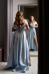 Young beautiful blonde girl wearing an off-the-shoulder full-length sky blue satin slit prom ball gown. Model looking in mirror. Fitting room in dress hire service.