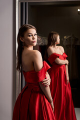 Young beautiful girl wearing an off-the-shoulder full-length crimson red satin slit prom ball gown. Model looking in mirror. Fitting room in dress hire service.