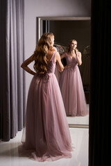 Young beautiful blonde girl wearing a full-length pink violet or purple chiffon prom ball gown decorated with sparkles and sequins. Model in front of mirror in a fitting room at dress hire service.