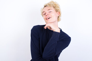 Optimistic Young handsome Caucasian blond man standing against white background keeps hands partly crossed and hand under chin, looks at camera with pleasure. Happy emotions concept.