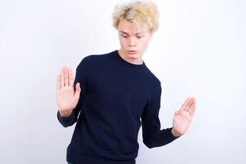 Serious Young handsome Caucasian blond man standing against white background pulls palms towards camera, makes stop gesture, asks to control your emotions and not be nervous