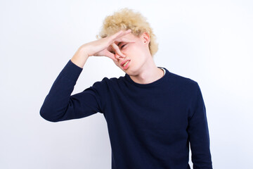 Young handsome Caucasian blond man standing against white background smelling something stinky and...