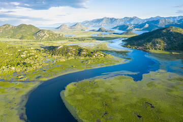 Blue river running through green valley toward distant mountains. Bends and curves of Crnojevica river in Montenegro, meandering through the marsh on its way to lake Skadar. Aerial view.