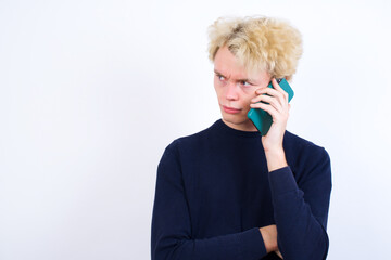 Sad Young handsome Caucasian blond man standing against white background  talking on smartphone. Communication concept.