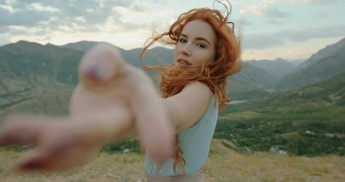 Beautiful female model on top of a mountain posing for photoshoot, reaching her hand out to camera while wind blows her dress and red hair - freedom, tranquility 4k footage