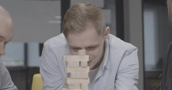 Close-up portrait of young handsome concentrated man playing Jenga with friends. Headshot of smiling positive Caucasian guy enjoying leisure game indoors. Cinema 4k ProRes HQ.