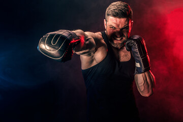 Fototapeta na wymiar Bearded tattooed sportsman muay thai boxer in black undershirt and boxing gloves fighting on dark background with smoke. Sport concept.
