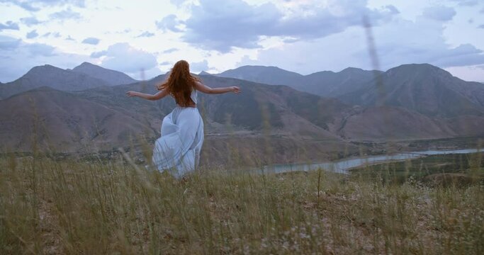 Young beautiful girl with red hair wearing white dress walking on top of a mountain facing wind blowing her hair and dress - freedom, adventure, harmony 4k footage