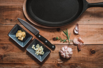 Iron pan, ready to cook, on a kitchen table, accompanied with butter, minced garlic and a kitchen knife, garlic and rosemary, on a rustic wooden background