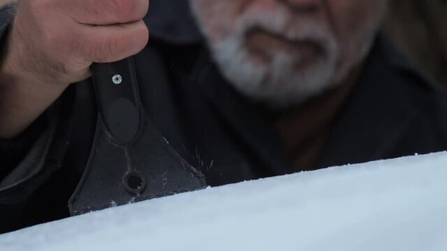 Aged businessman with grey beard cleans electric car roof with black and yellow snow brush on cold day