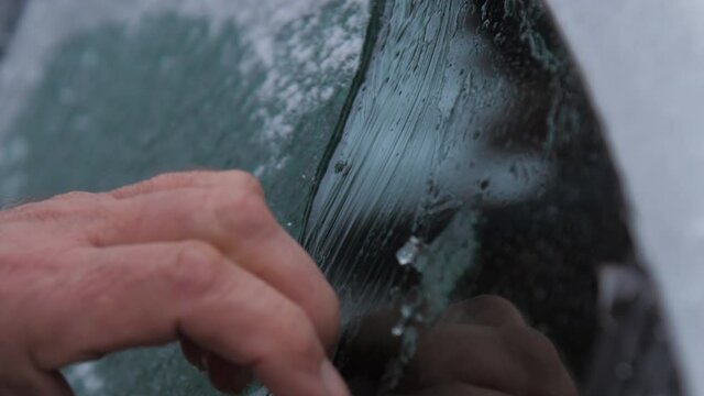 Person cleans electric car window with hands and black rubber ice scraper on cold winter day. Concept harsh weather conditions