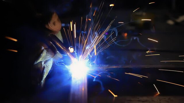 Selective focus of sparks of electric grinding machine used by young woman wearing safety gloves in workshop to join corners of rod