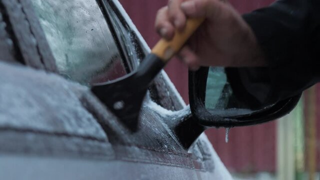 Person hand holds black and yellow ice scraper and cleans electric vehicle door on colds winter day. Concept weather conditions