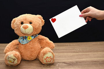 A teddy bear is on the table and the girl holds out a white envelope with a heart. Valentine's Day gift
