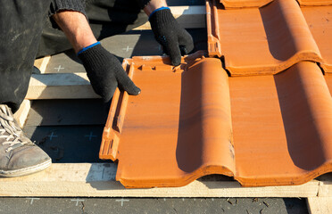 Closeup of worker hands installing yellow ceramic roofing tiles mounted on wooden boards covering...