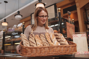 Low angle shot of a mature woman holding basket full of freshly baked bread, working at her bakery store